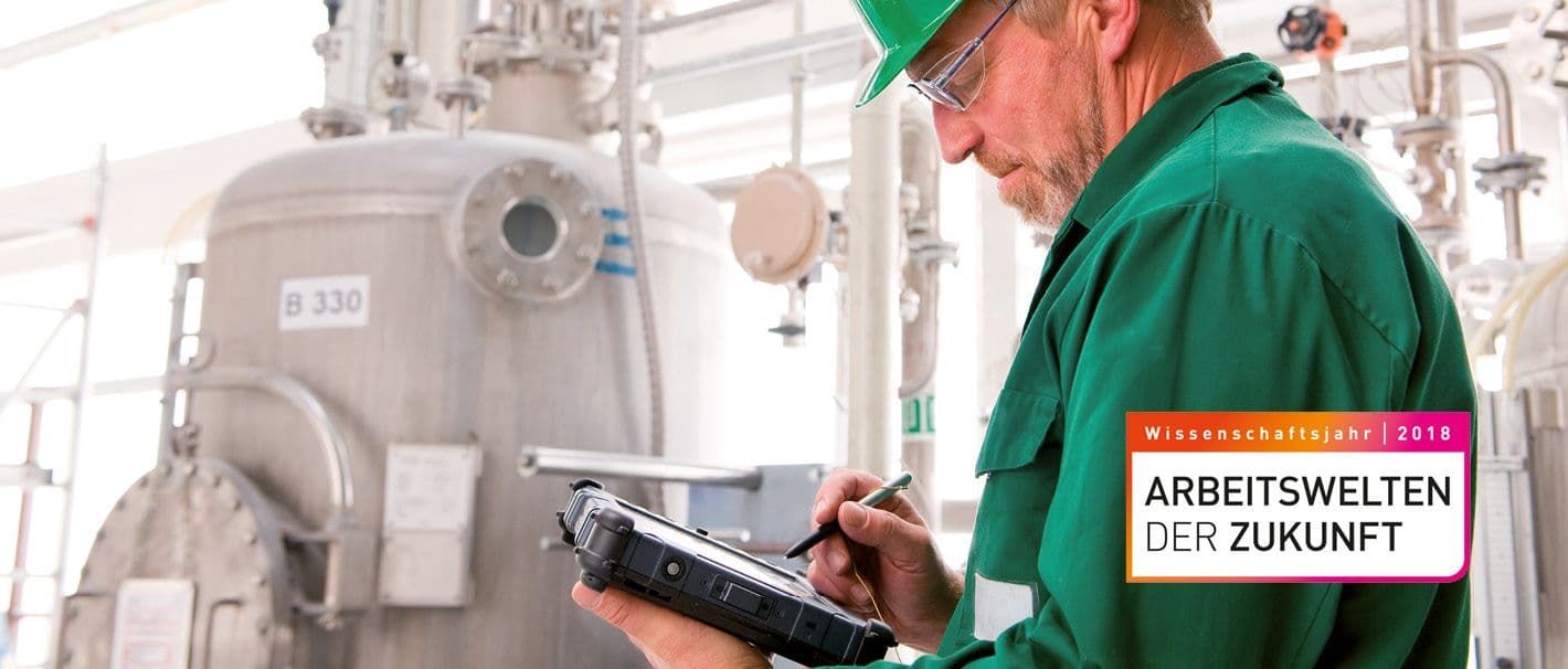 Älterer Herr mit grünem Arbeitsanzug hält elektronischen Stift über einem Tablet vor einem großen Kessel in einer Fabrikhalle.