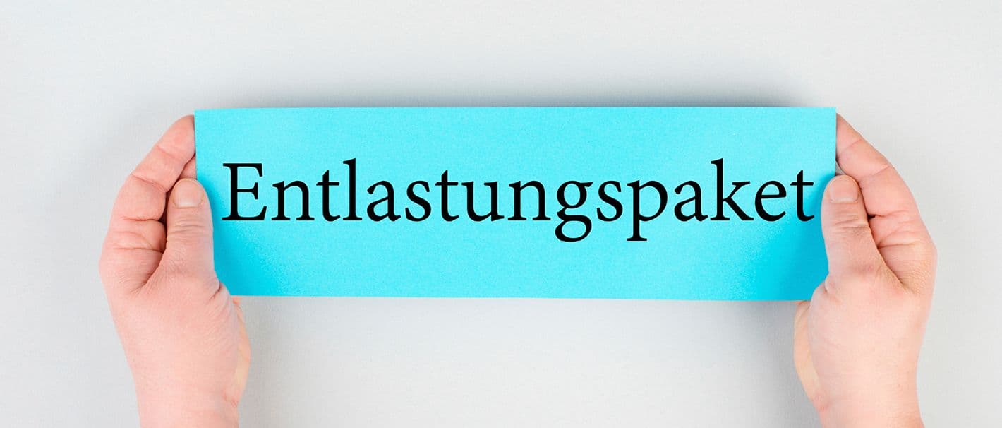 Zwei Hände halten einen blauen Zettel, auf dem „Entlastungspaket“ steht.