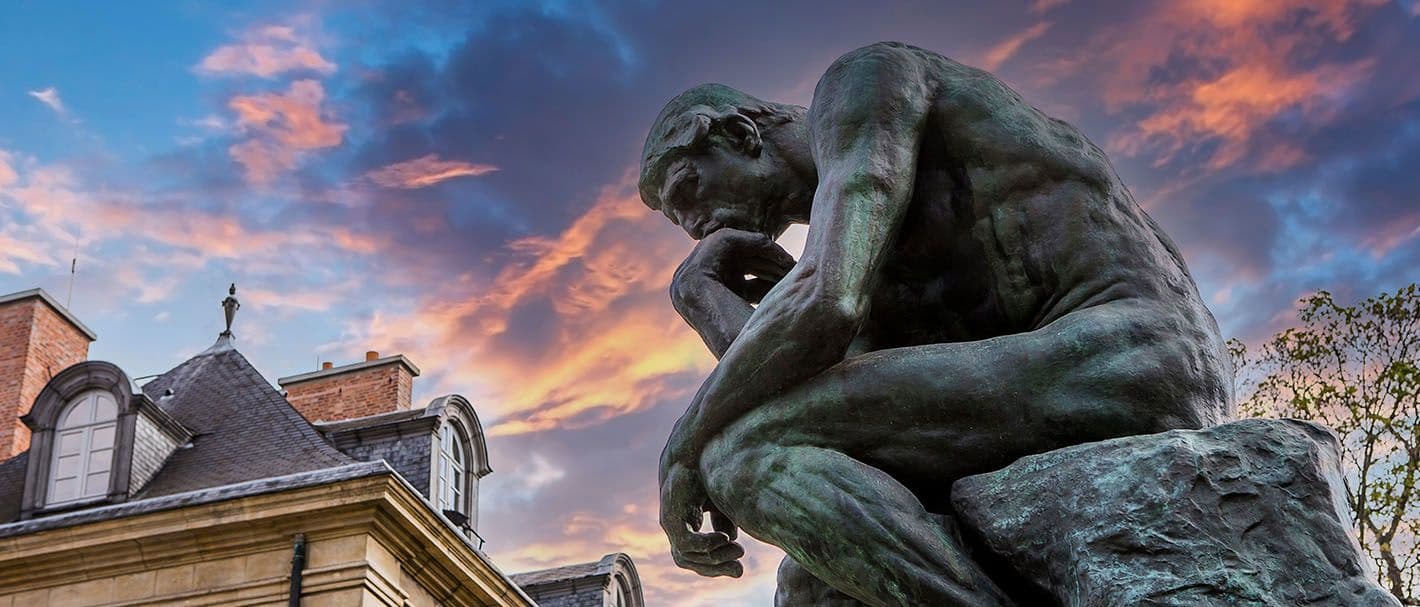 Das Foto zeigt Augustin Rodins Plastik “Der Denker” im Garten des Musée Rodin in Paris. Das Foto wurde seitlich und aus der Froschperspektive aufgenommen, vor einem Himmel mit orangenen und graublauen Wolken und einem Gebäude.