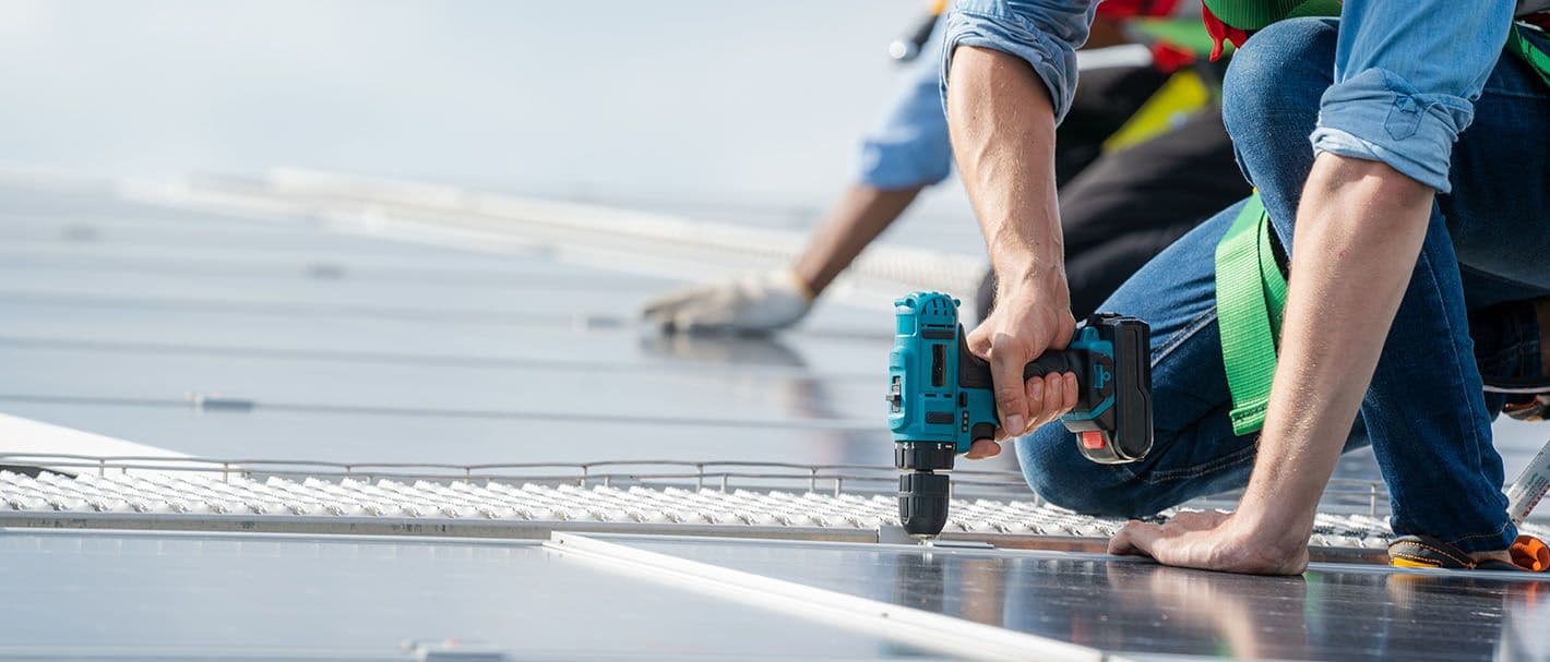 Ein Handwerker installiert mit einer Bohrmaschine Solarpanele auf einem Gebäudedach.