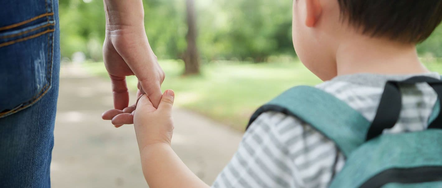 Aufnahme von hinten: Eine Mutter und ihr kleiner Sohn laufen Hand in Hand. Der Sohn trägt einen Rucksack.