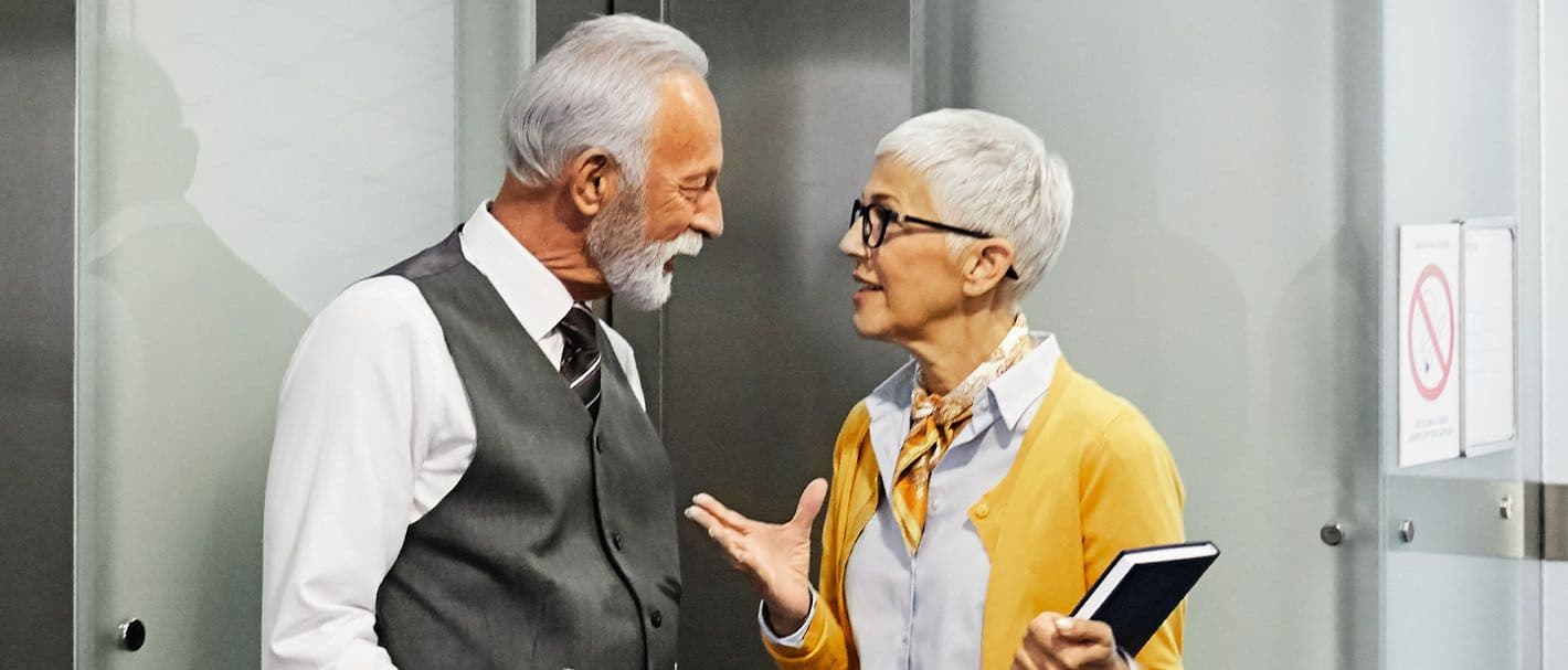 Zwei ältere Geschäftsleute, ein Mann und eine Frau, stehen in einem Bürogebäude vor einem Aufzug und unterhalten sich.
