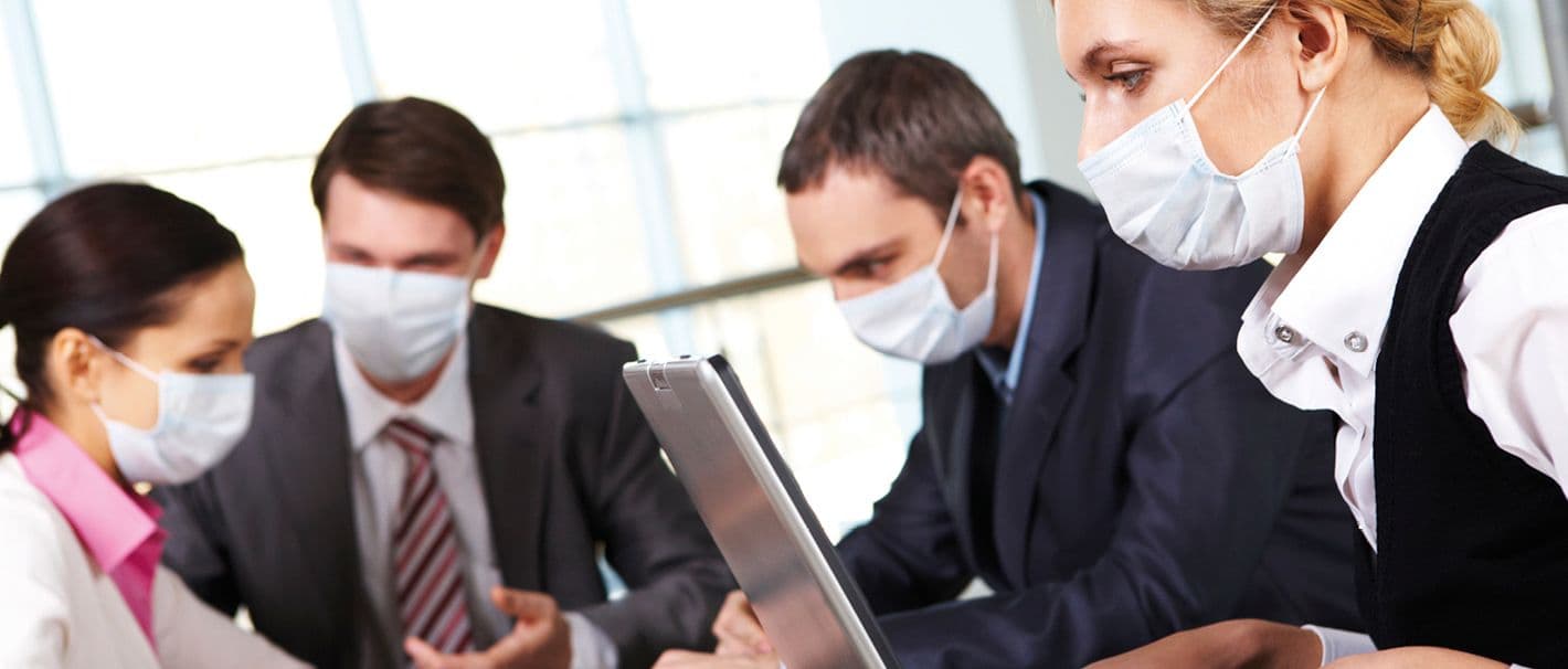 Gruppe von Beschäftigten, die um einen Arbeitstisch herumsitzen und Mundschutz tragen