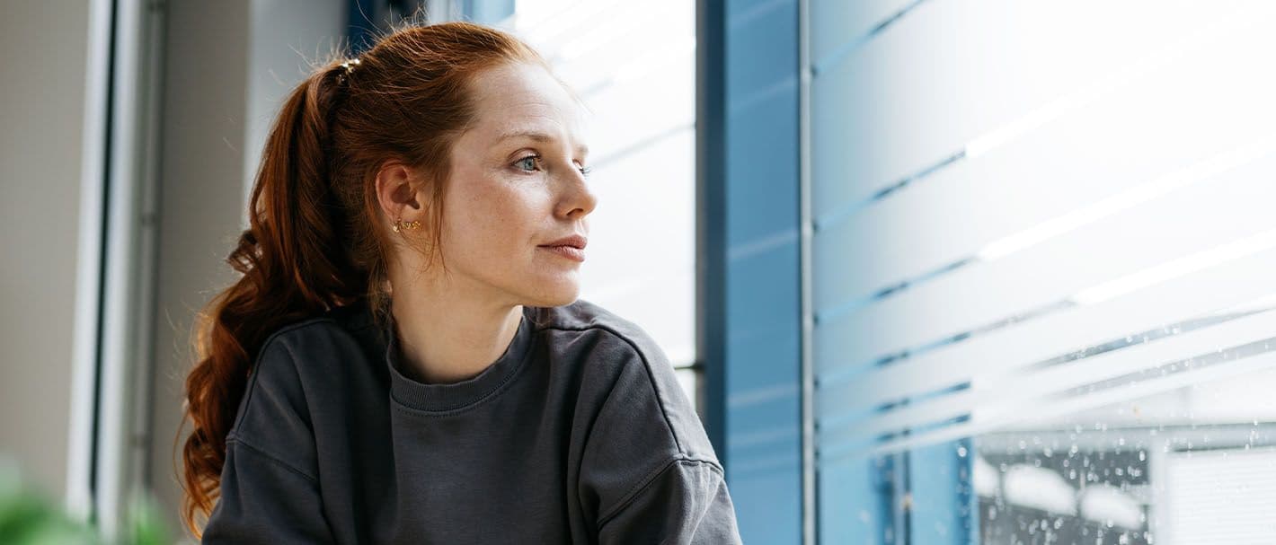 Eine junge Frau sitzt an einem Tisch am Fenster und schaut gedankenverloren hinaus. Auf dem Tisch befinden sich eine Tasse, ein Handy und eine Topfpflanze.