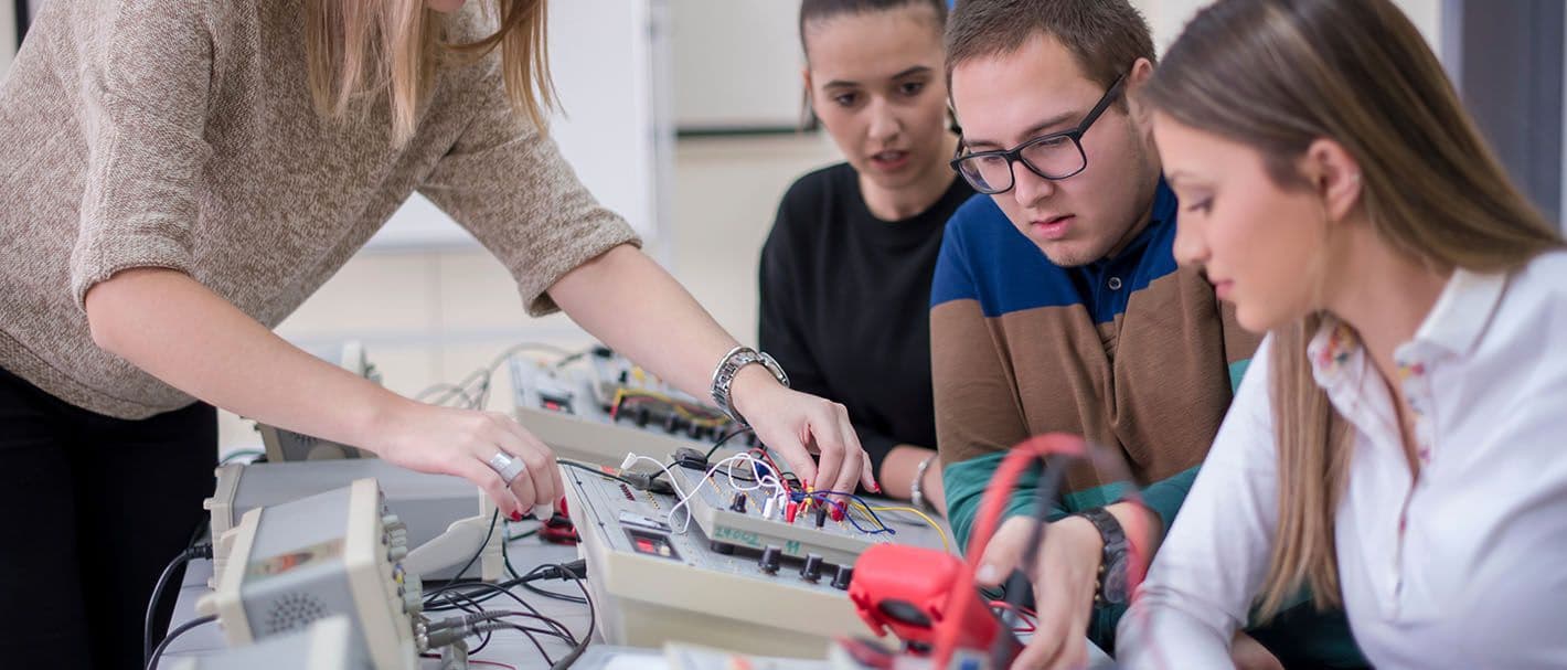 Eine Frau nutzt elektrotechnische Geräte und zwei junge Frauen und ein junger Mann sehen dabei zu