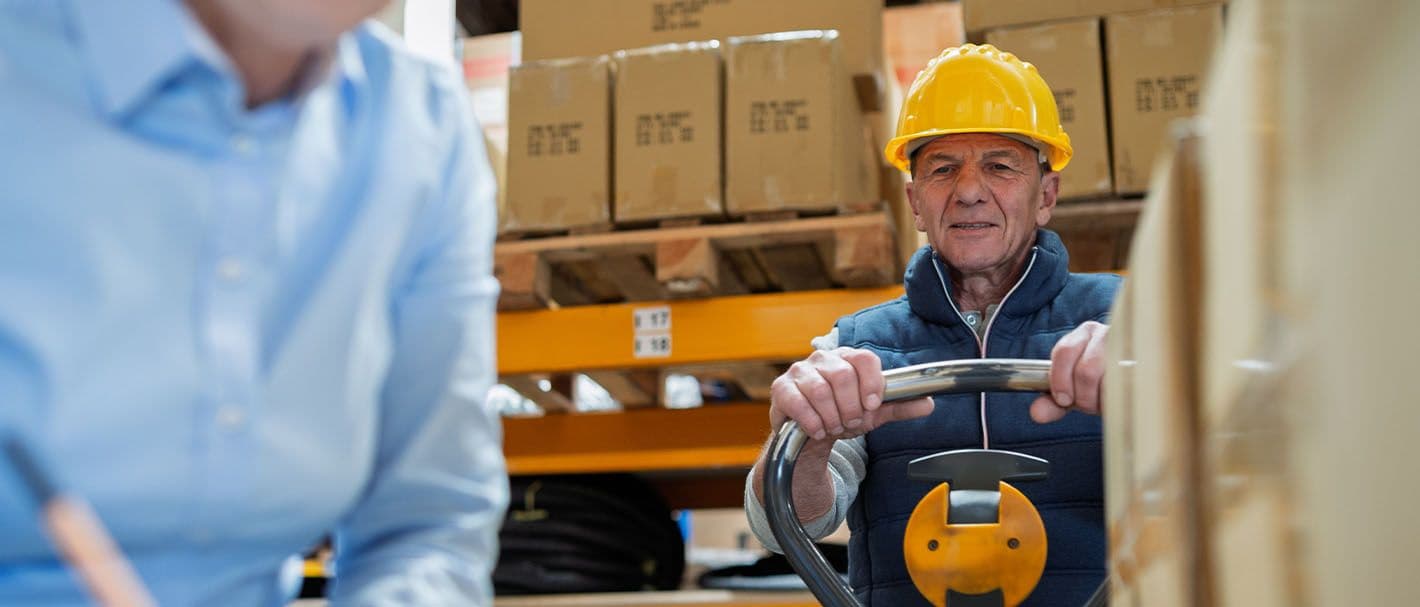 In einem Warenlager beobachtet ein älterer Arbeiter an einem Handhubwagen eine leitende Angestellte, die etwas auf einen Schreibblock notiert. Der Fokus liegt auf dem Arbeiter.