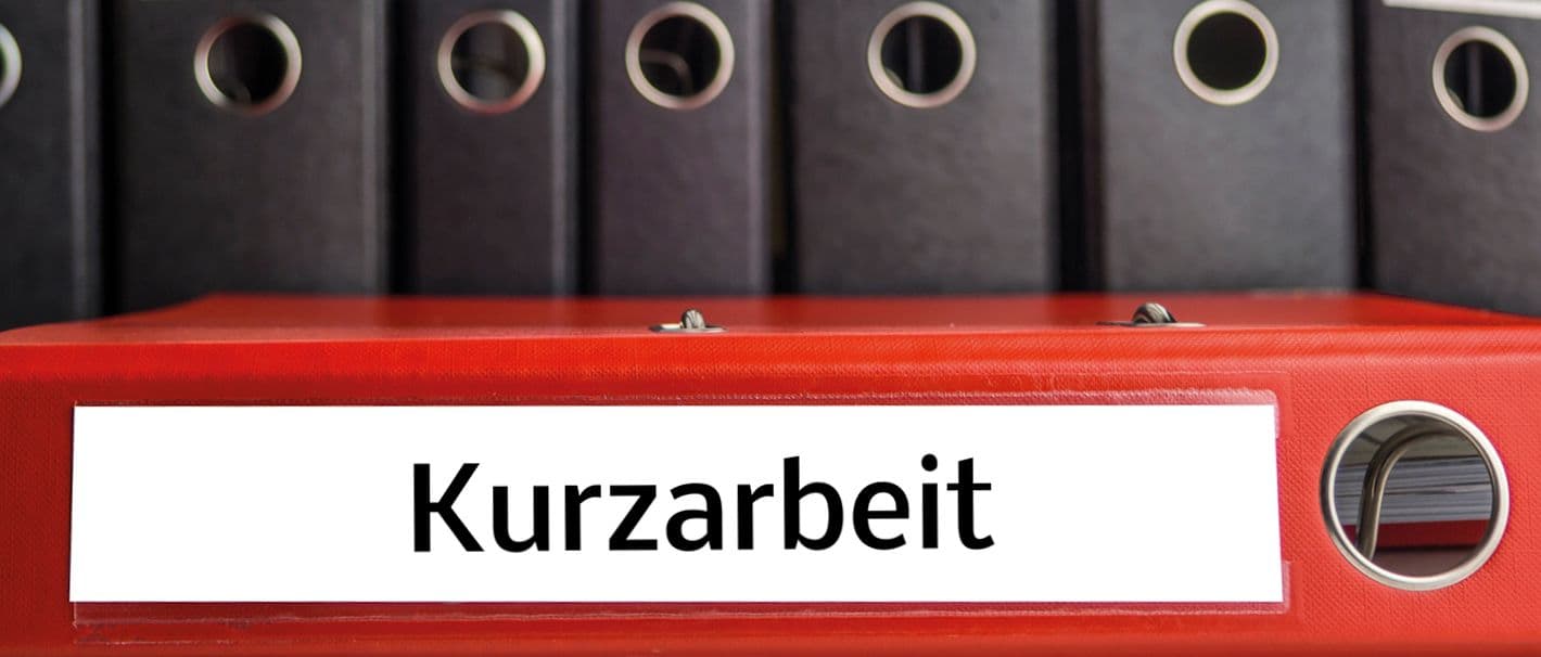 Aktenordner mit der Aufschrift „Kurzarbeit“ liegt quer im Bild. Im Hintergrund sind stehen schwarze Aktenordner aufrecht da.