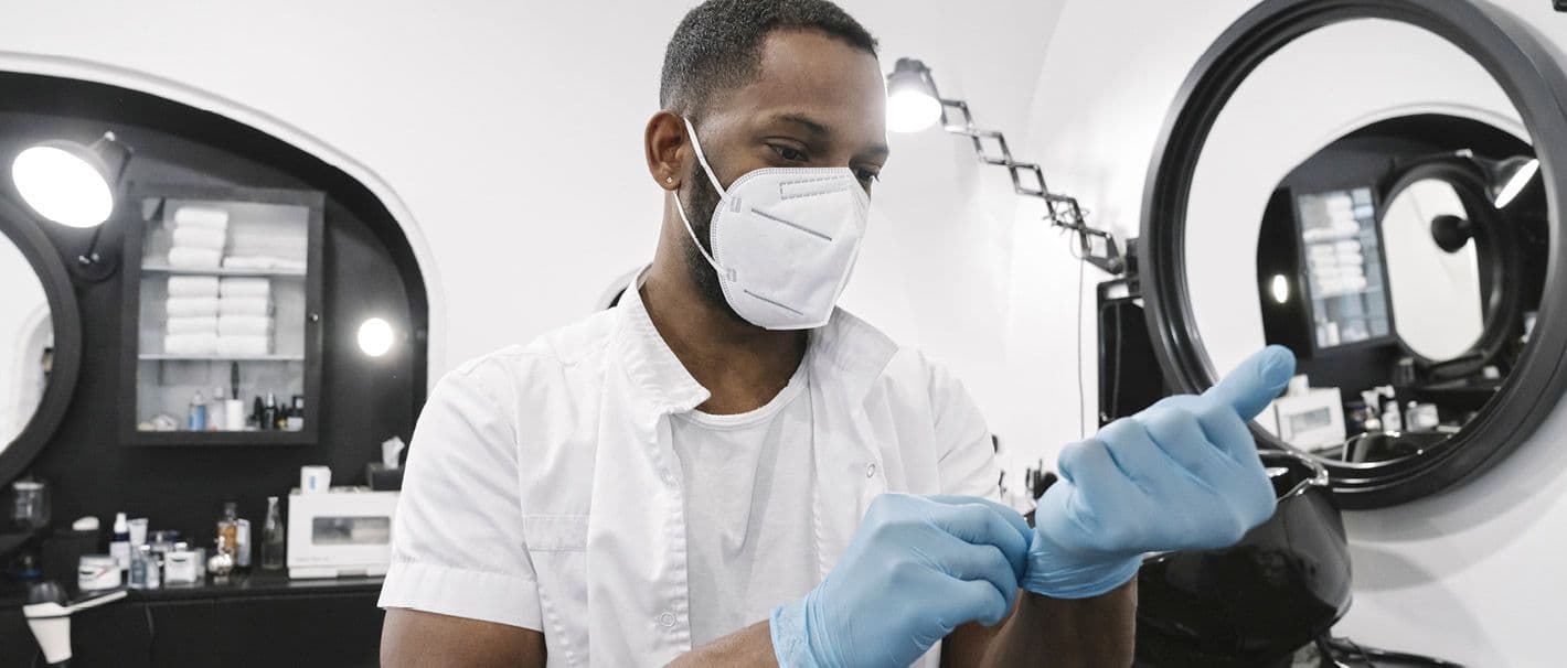 Friseur mit Migrationshintergrund und Mundschutz zieht sich in seinem Friseursalon Einmalhandschuhe an