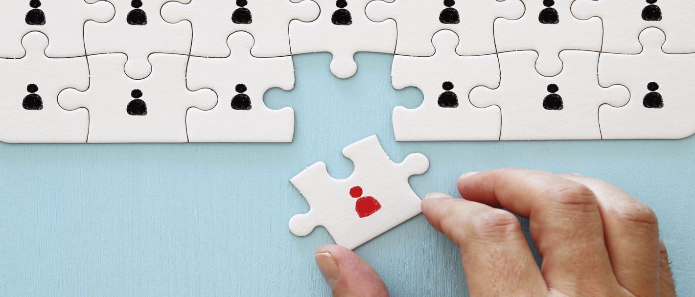 Eine Hand fügt ein Puzzlestück mit einer roten Figur in ein Puzzle mit vielen schwarzen Figuren ein.