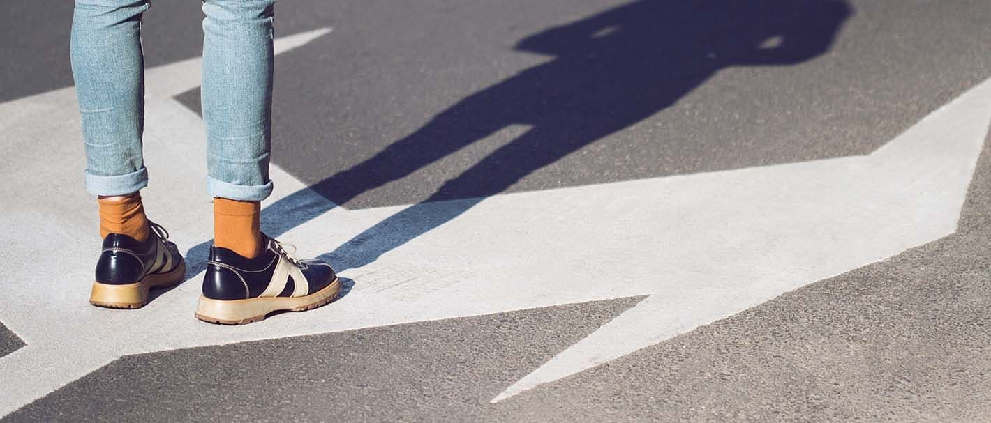 Nahaufnahme der Beine und Füße einer Frau, die auf der Straße vor zwei Pfeilen steht. Einer zeigt nach links und einer nach rechts.