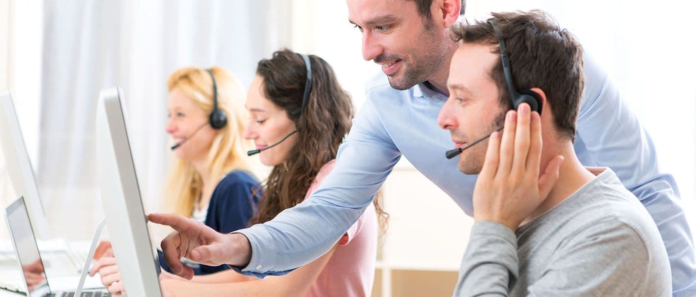 Schulungssituation für Callcenter-Mitarbeiter: zwei Frauen und ein Mann sitzen mit Headsets vor ihren Computern. Ein Mann im Hemd steht hinter dem Mann mit dem Headset und zeigt auf dessen Monitor. Alle Personen schauen freundlich.