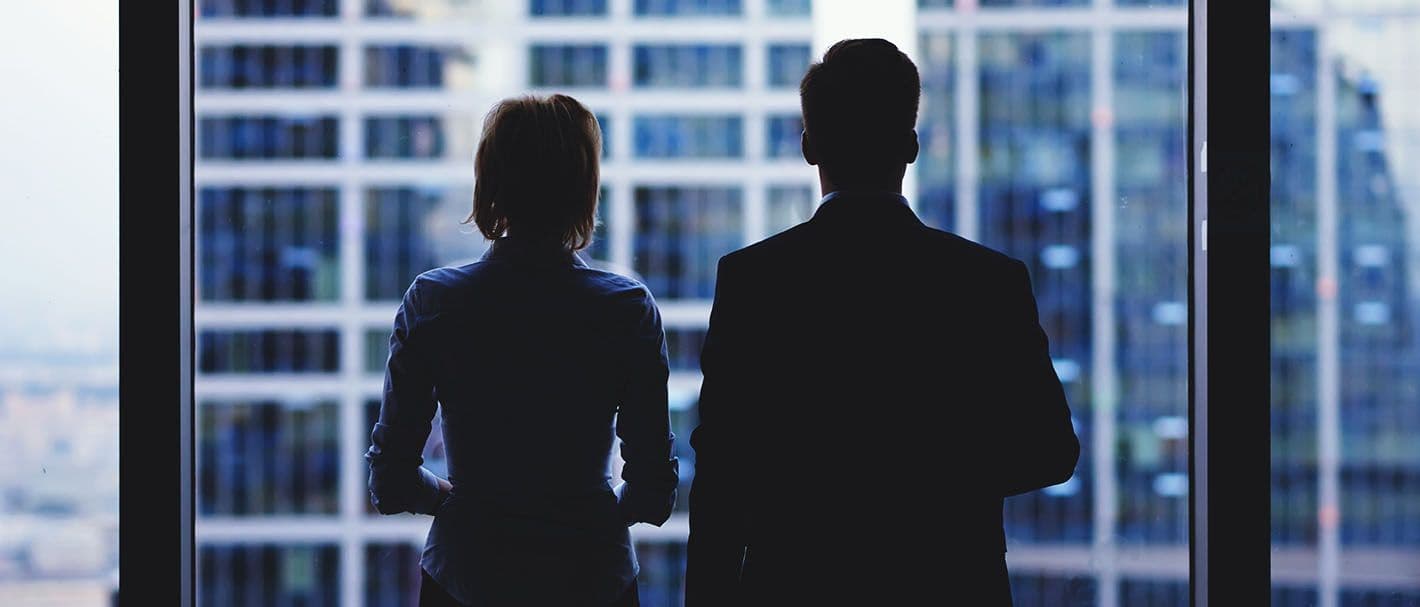 Zwei Geschäftsleute, eine Frau und ein Mann schauen aus dem Fenster eines Bürogebäudes, wobei im Gegenlicht nur deren Silhouetten zu erkennen sind.