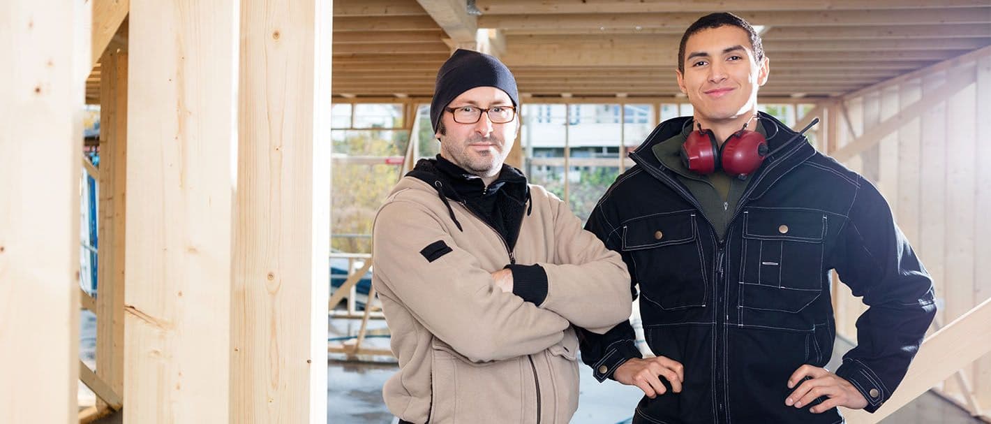Portrait von zwei Zimmerern, die selbstbewusst auf ihrer Baustelle stehen und zum Betrachter blicken.