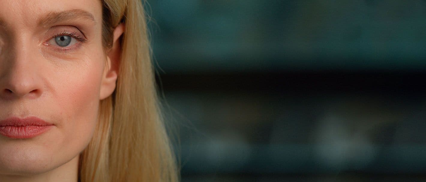 Frontale Aufnahme des Gesichts einer erwachsenen Frau mittleren Alters mit heller Haut, blondem Haar und grünblauen Augen. Sie sieht den Betrachter mit einem neutralen Gesichtsausdruck direkt an. Das Bild ist angeschnitten, es ist nur die Hälfte des Gesichts abgebildet.