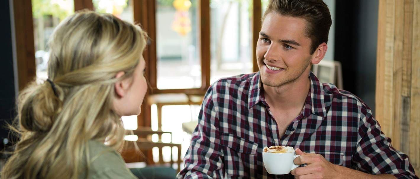 Ein Junger Mann mit eine Kaffeetasse in der Hand ist im Gespräch mit einer jungen Frau. Das Bild steht für die Jobvermittlung im Cafe.