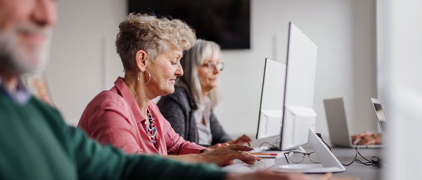 Perspektivische Aufnahme von erwachsenen Kursteilnehmern, die nebeneinander in einer Reihe an Computern arbeiten. Im Fokus ist eine Frau, die in der Mitte sitzt.