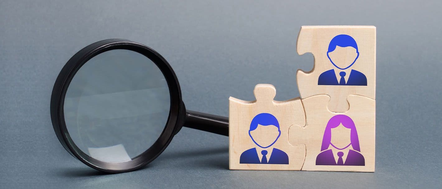 Three interlocking puzzle pieces, each with a symbol for a person, are in front of a magnifying glass.