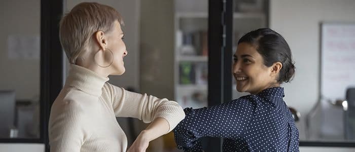 Zwei Frauen berühren sich gegenseitig mit ihren Ellbogen und lächeln sich an.