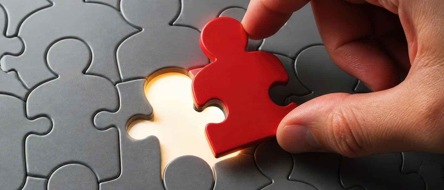 Hand, die ein rotes Puzzleteil in Form einer Person in eine passende Lücke in ein Puzzle einsetzt, das aus grauen Personen-Teilen besteht.