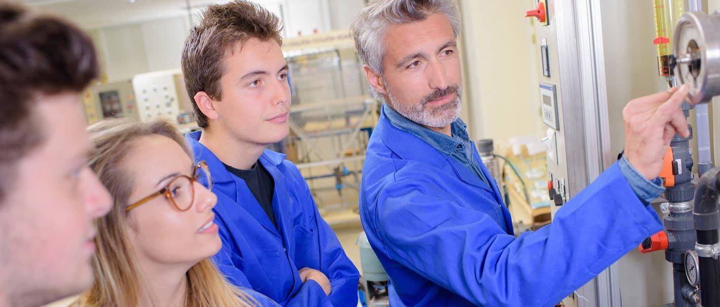 Drei Auszubildende in blauen Anzügen folgen den Erklärungen ihres Ausbilders.