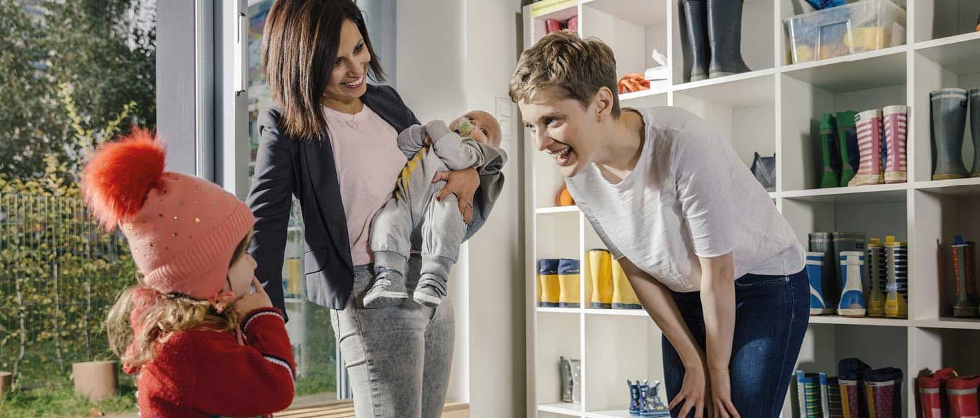 Mutter mit zwei kleinen Kindern wird in der Kita von einer Erzieherin begrüßt