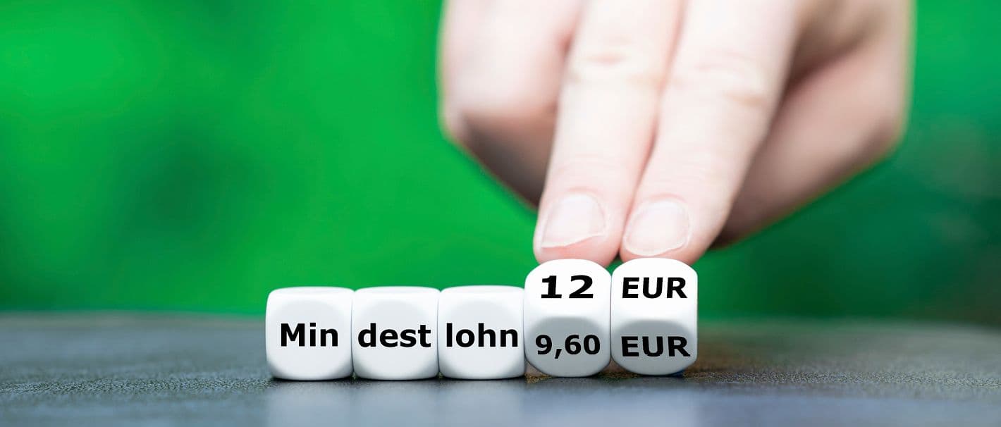 5 weiße Würfel bilden zusammen das Wort Mindestlohn. Eine menschliche Hand dreht die beiden letzten Würfel von 9,60 auf 12 Euro.