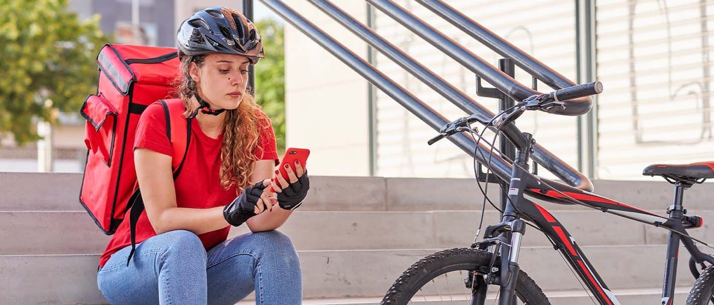 Fahrradkurierin sitzt neben ihrem Fahrrad auf einer Treppe und überprüft auf ihrem Handy, ob neue Aufträge vorliegen.