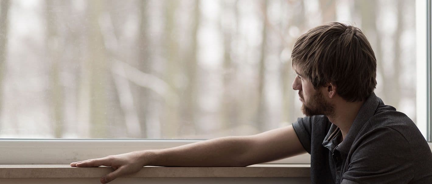 Jüngerer Mann sitzt am Fenster uns schaut hinaus auf kahle Baumstämme bei trübem Wetter.
